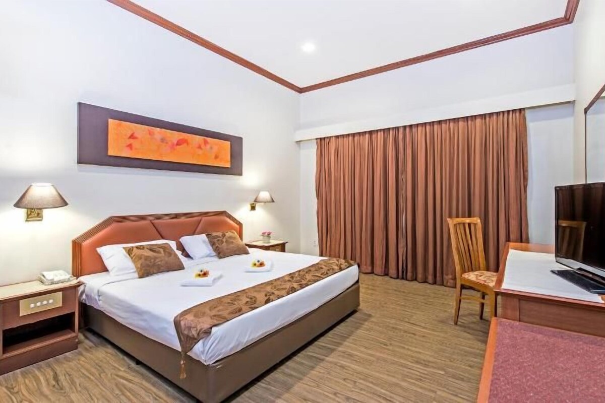Una habitación de hotel con una cama grande, dos lámparas de noche, un escritorio con televisor, una silla y un ventanal con cortinas.