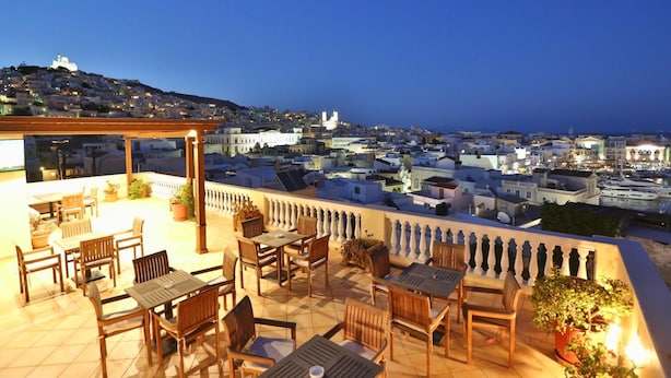 Una terraza con muebles de madera y plantas en macetas, con vistas a un paisaje urbano con edificios y una estructura en la colina.