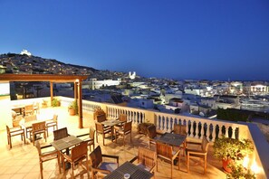 A terrace with wooden furniture and potted plants, overlooking a cityscape with buildings and a hilltop structure. - Aigli Hotel Syros (Syros)