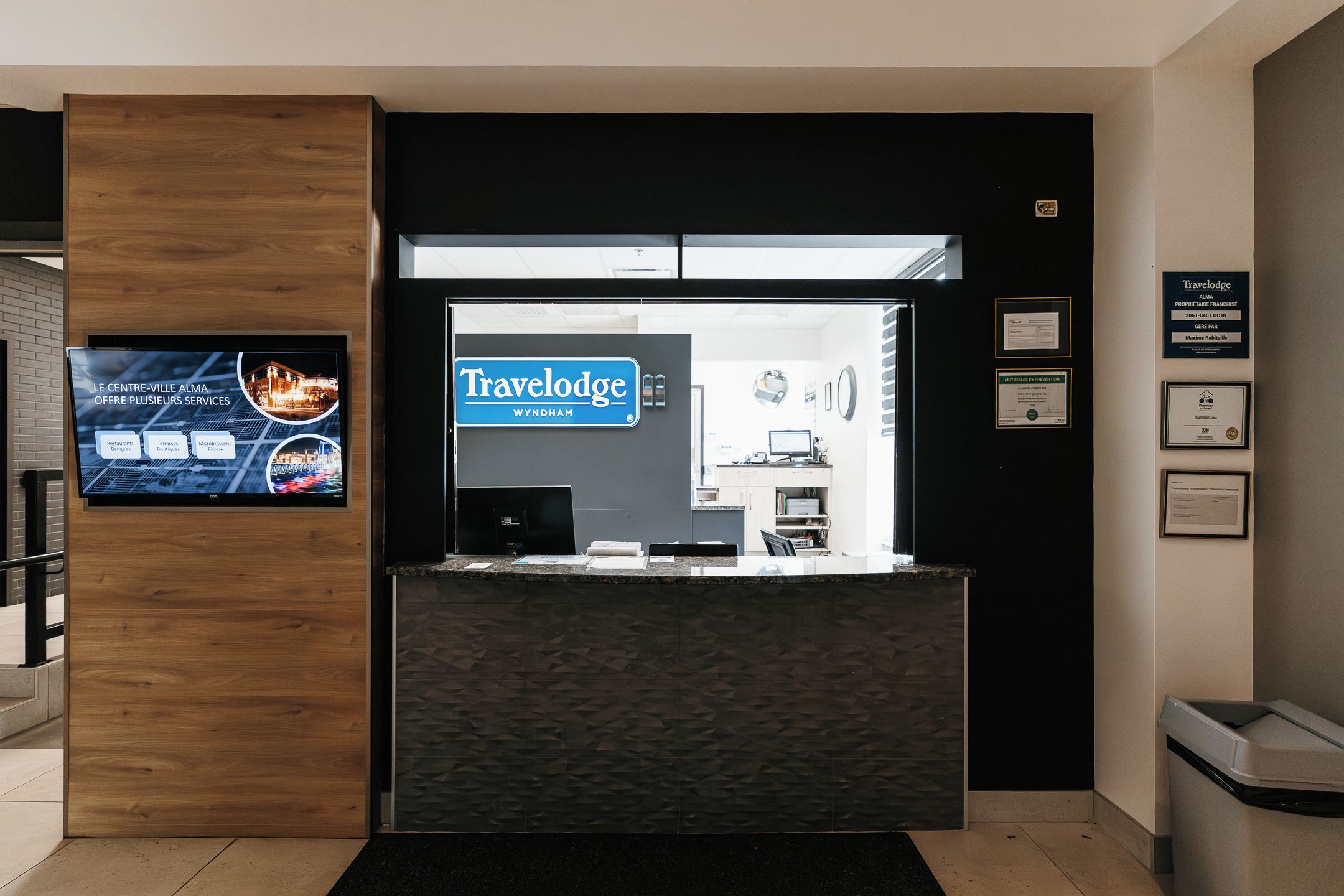 Una recepción con un cartel de Travelodge Wyndham, una pantalla digital mostrando atracciones locales y certificados enmarcados en la pared.