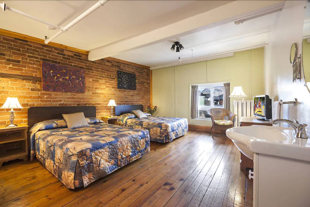 Une chambre avec deux lits, un plancher en bois, un mur en briques, une grande fenêtre et un lavabo.