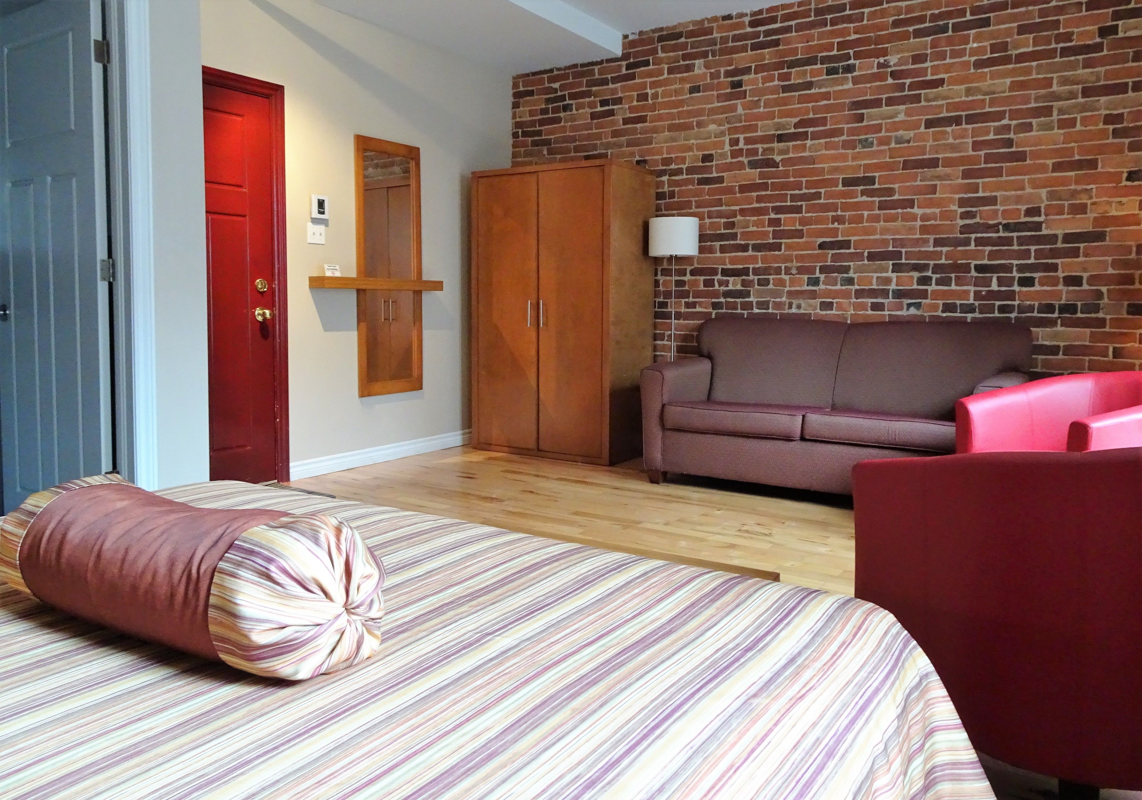 Una habitación con una cama a rayas, una puerta roja, una pared de ladrillo, un armario de madera, un sofá rosa y una silla roja.
