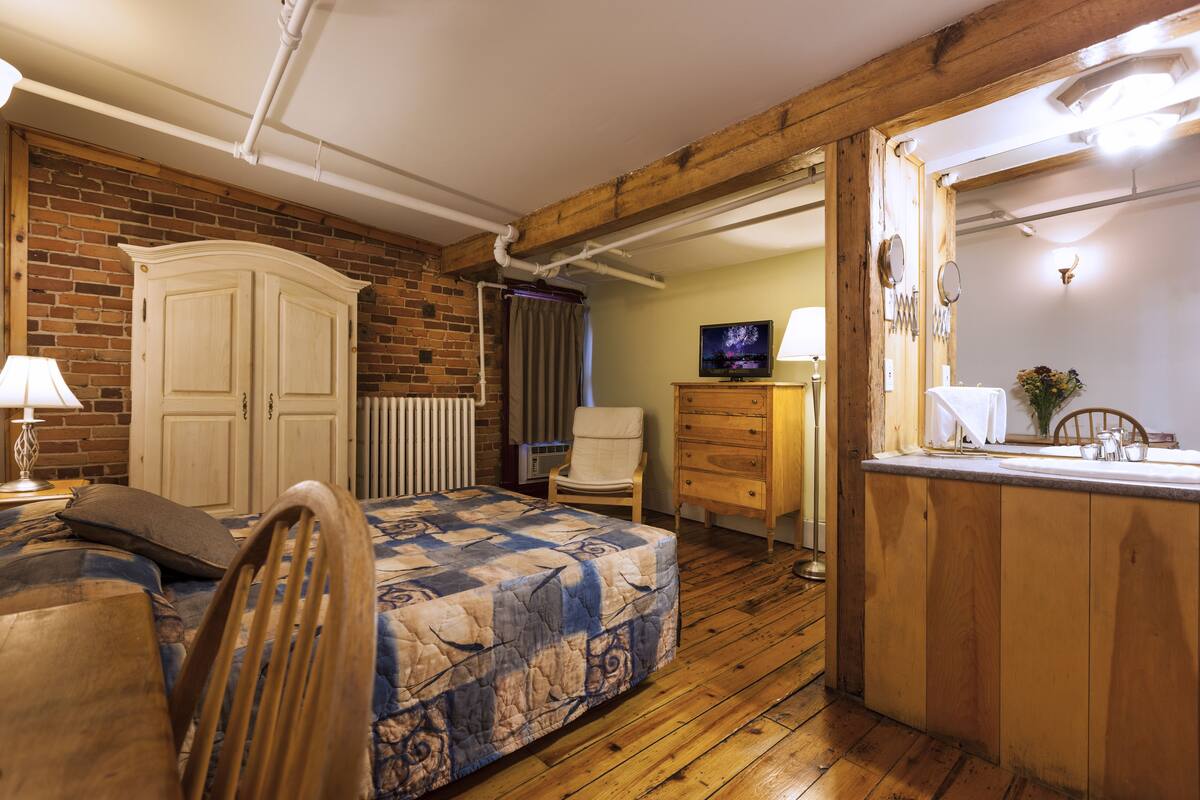 Une chambre avec un plancher en bois, un lit avec une couette, une chaise, une commode, un téléviseur et une salle de bain avec un miroir et un lavabo.