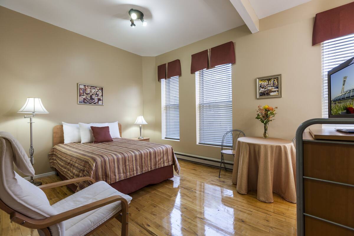 Une chambre d’hôtel comprenant un lit, des lampes de chevet, une chaise, une petite table avec un vase et deux fenêtres avec des rideaux.