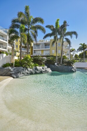 A clear pool surrounded by palm trees and a rocky edge, with a building in the background. - Seachange Coolum Beach (Coolum Beach)