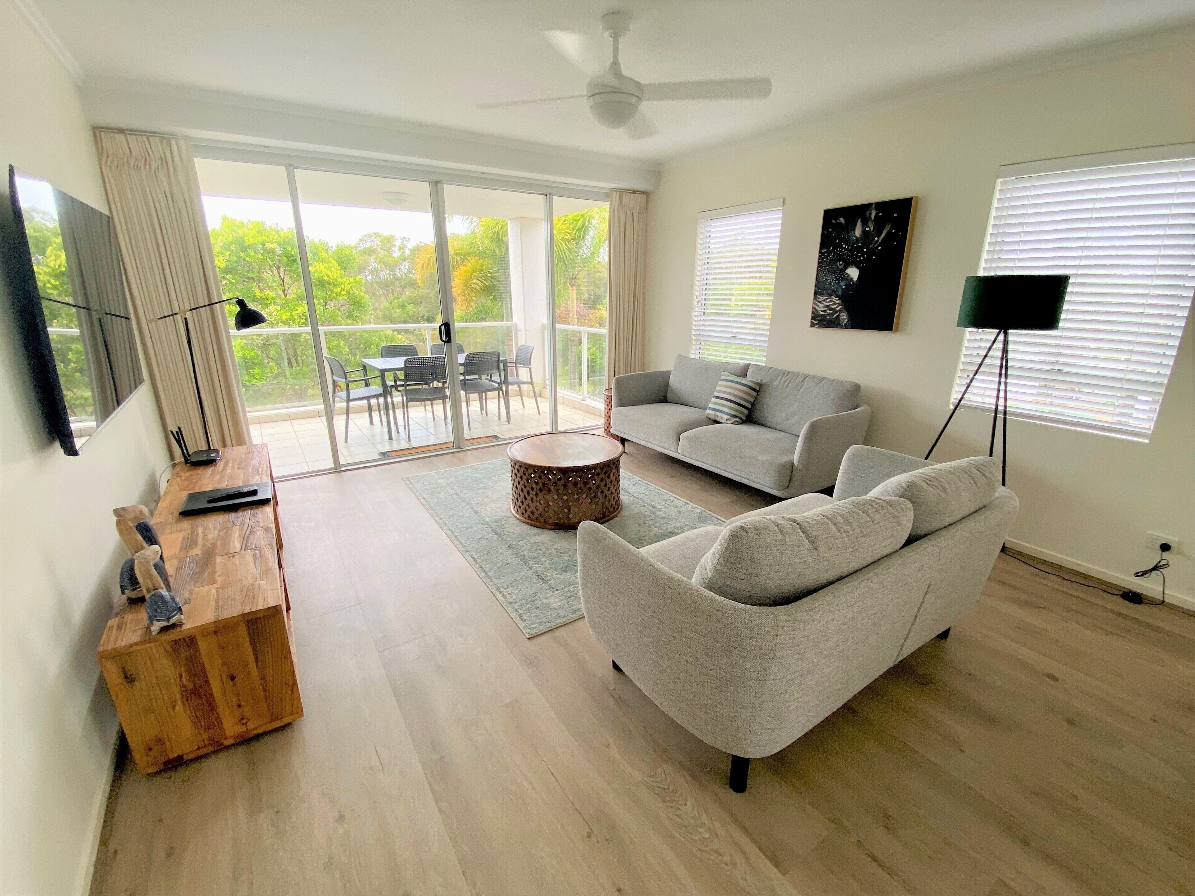 A spacious living room with a grey sofa, a wooden coffee table, and a TV on a wooden stand. There is a balcony with a glass door and a ceiling fan.