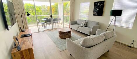 A spacious living room with a grey sofa, a wooden coffee table, and a TV on a wooden stand. There is a balcony with a glass door and a ceiling fan.
