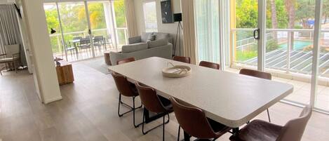 A spacious dining area with a long table and six chairs, large windows offering a view of a pool and greenery, and a modern kitchen in the background.