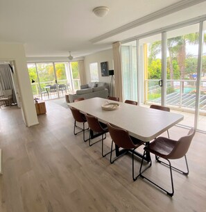 A spacious dining area with a long table and six chairs, large windows offering a view of a pool and greenery, and a modern kitchen in the background. - Seachange Coolum Beach (Coolum Beach)