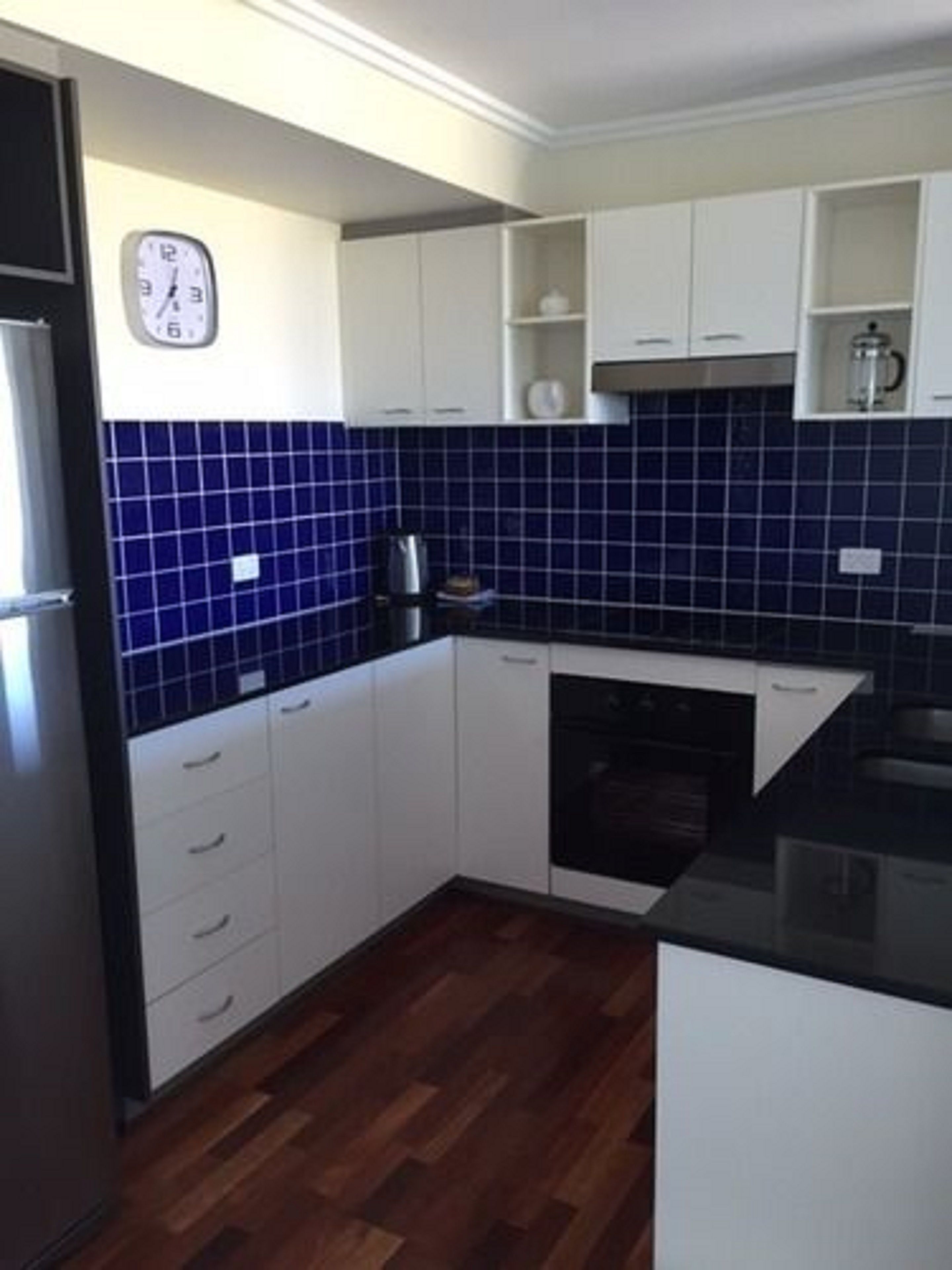 A kitchen with white cabinets, a blue tiled backsplash, a clock on the wall, and a coffee maker on the counter.