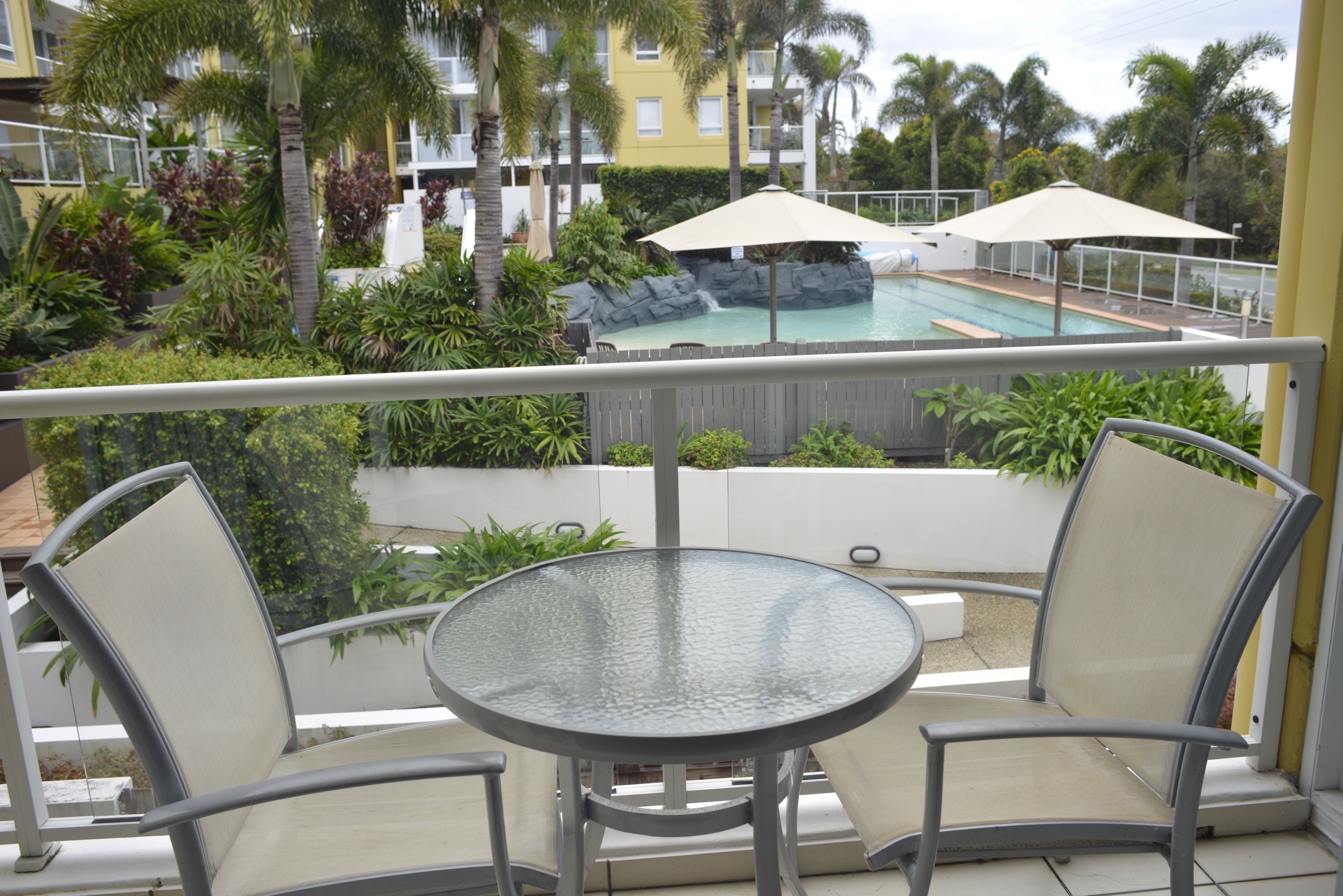 Un balcon doté d’une table en verre et de deux chaises, donnant sur un espace piscine agrémenté de parasols et d’une végétation luxuriante.
