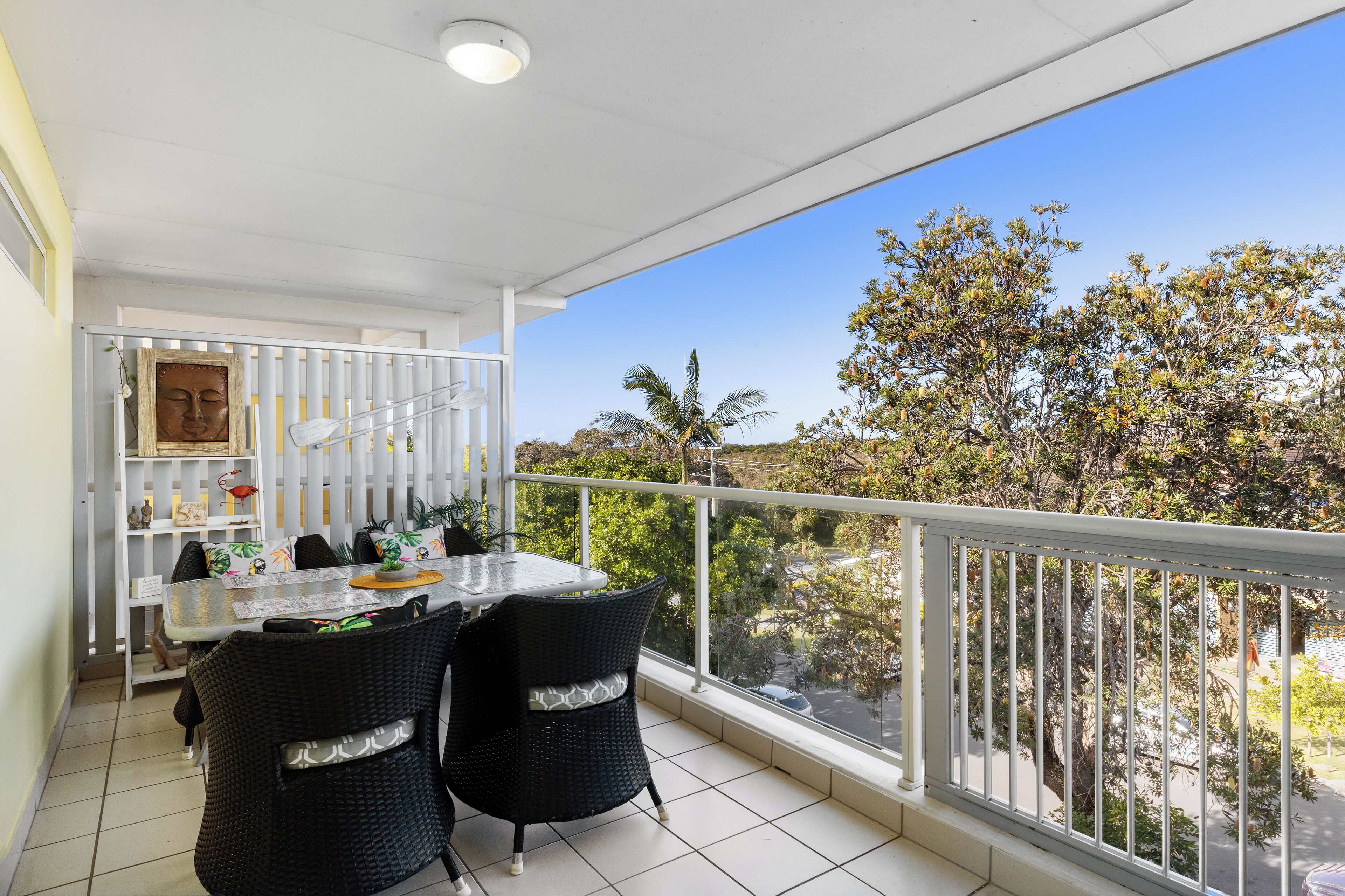 A balcony with a dining area, white railing, and a view of trees and a road.