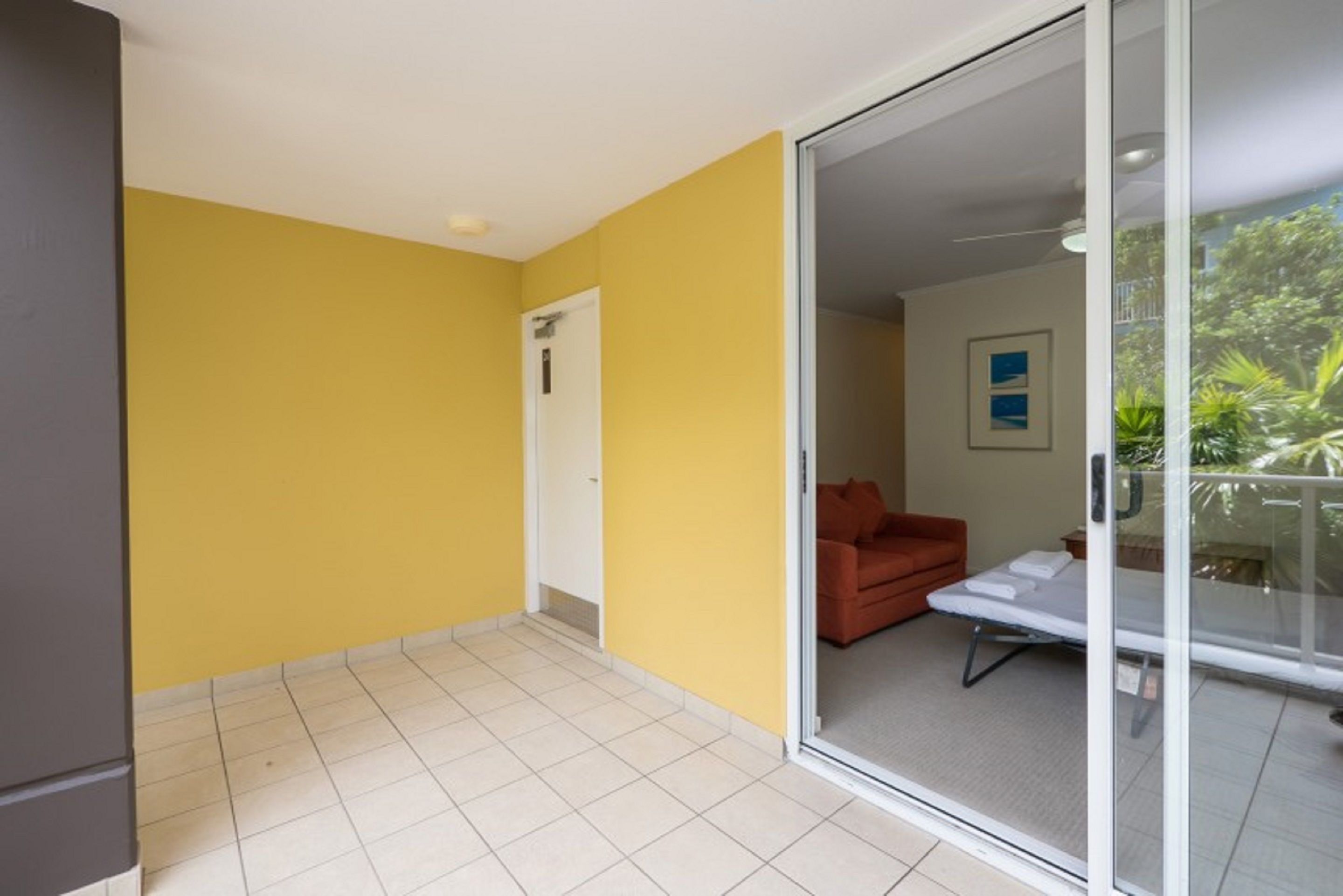 A room with yellow walls, a sliding glass door leading to a balcony with a red sofa and a mattress, and a white door.
