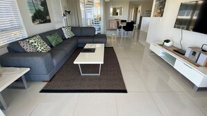 Un salon moderne avec un canapé d’angle gris, une table basse blanche et un tapis noir et blanc. On aperçoit à l’arrière-plan un coin cuisine avec une table et des chaises.
