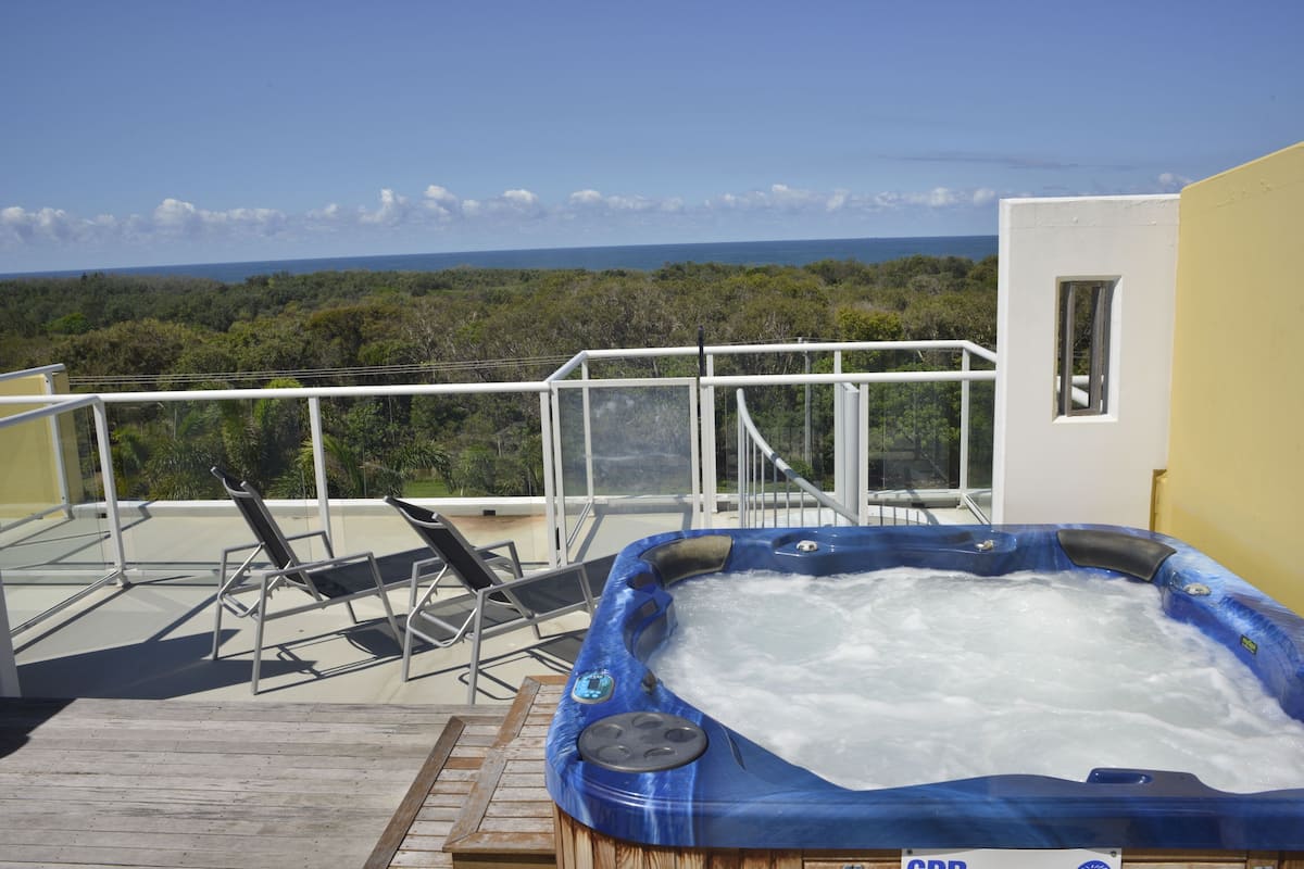 Un bain à remous sur une terrasse en bois, avec vue sur l’océan et les arbres.