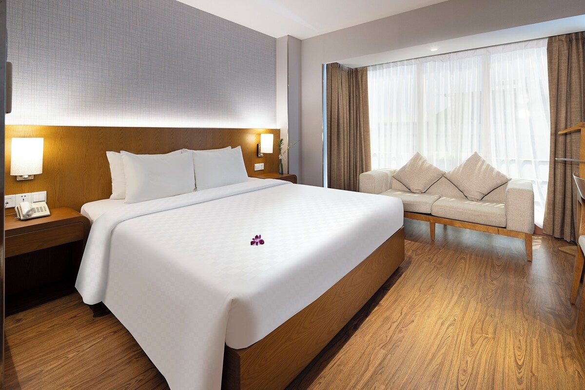 Een hotelkamer met een groot bed, een bank, een houten hoofdbord, een nachtkastje met een lamp en een raam met gordijnen.