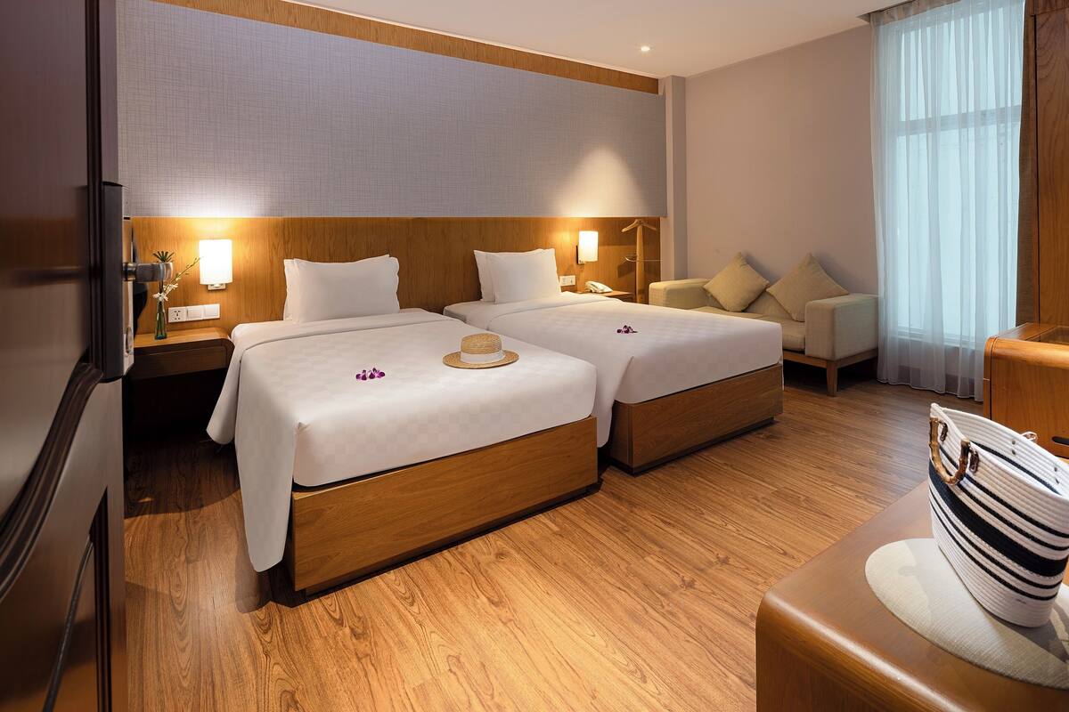 Quarto de hotel com duas camas, cabeceiras de madeira, mesas de cabeceira com luminárias, um sofá e uma grande janela com cortinas leves.
