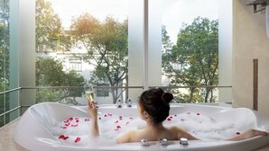 A woman in a bathtub filled with bubbles and rose petals, holding a glass of wine, with a view of trees and buildings outside.