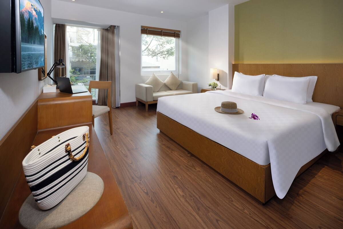 Quarto de hotel com uma cama grande, uma escrivaninha com um laptop, uma cadeira, um sofá e vista para o exterior.