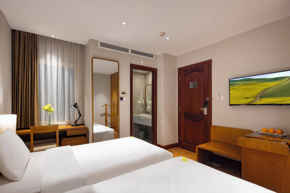 Quarto de hotel com duas camas, uma escrivaninha, um espelho, uma televisão e um banco de madeira.
