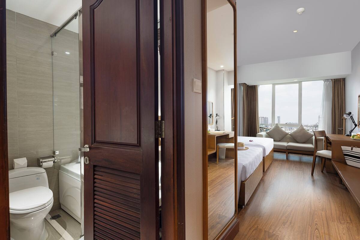 Quarto de hotel com porta de madeira, banheiro com vaso sanitário e pia, cama com mesa de cabeceira, escrivaninha com cadeira e uma janela grande com vista para a cidade.