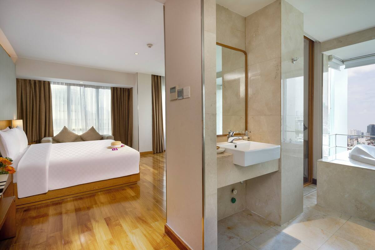 Quarto de hotel moderno com uma cama grande, piso de madeira, banheiro com pia e espelho, e vista da cidade pela janela.