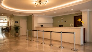 Zona de recepción de un hotel con un mostrador, una planta, un reloj y un cartel que dice 'AVISO'.