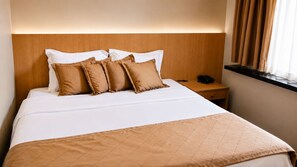 Habitación de hotel con cama, cabecera de madera, mesita de noche con teléfono y ventana con cortinas.