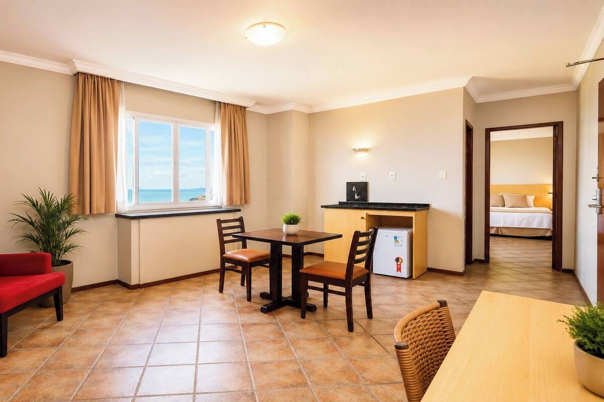 A compact hotel room with a dining area, a small kitchenette, a bed, and a view of the sea through a window.