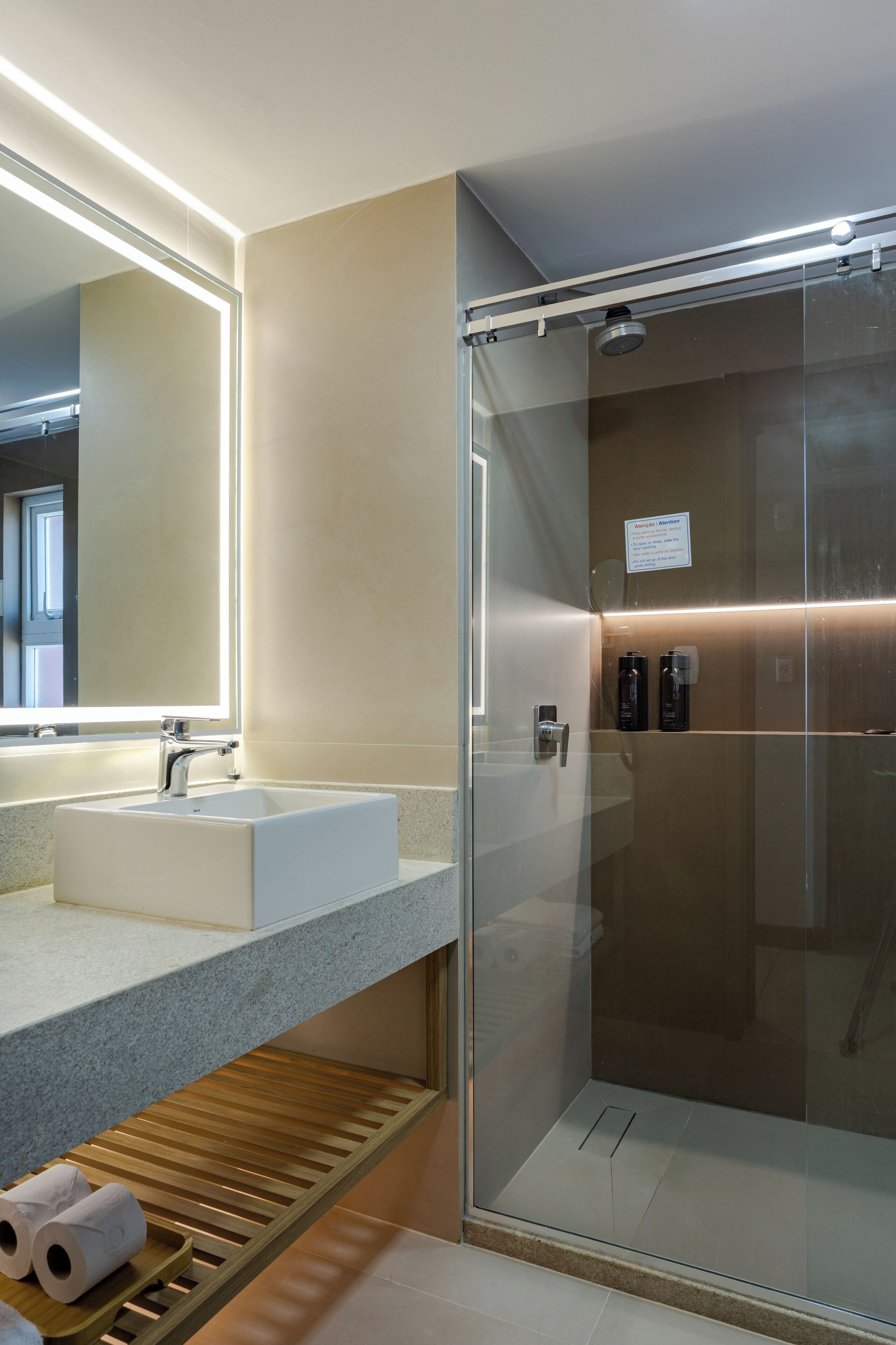 Une salle de bain moderne avec une douche à parois de verre, un lavabo avec un miroir au-dessus et une étagère en bois où sont rangés les articles de toilette.