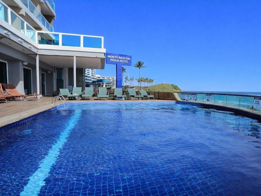 A swimming pool with lounge chairs, a sign for Monte Pascoal Praia Hotel, and a view of the ocean.