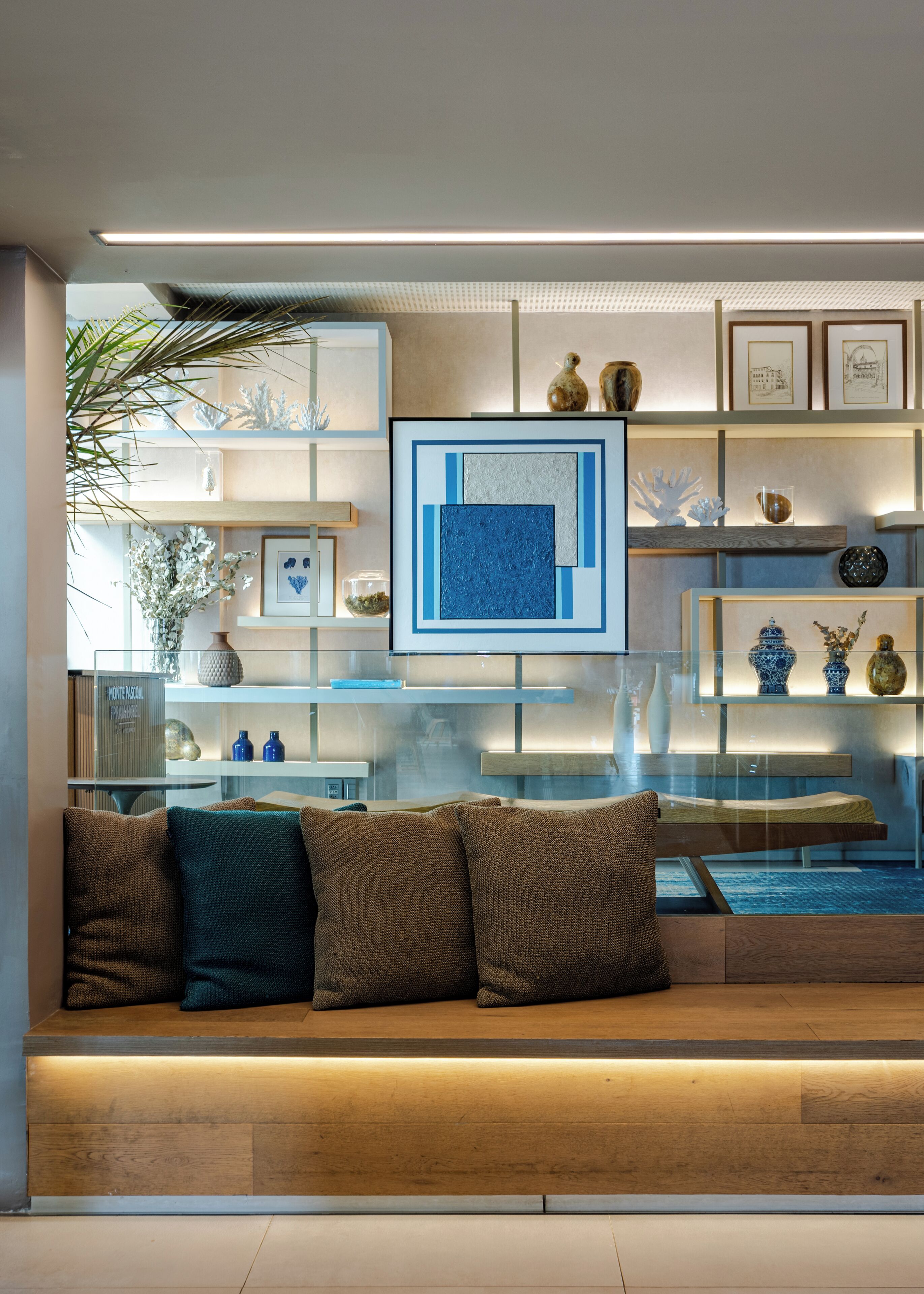 A modern living room with a glass partition, wooden shelves, and a cushioned bench.