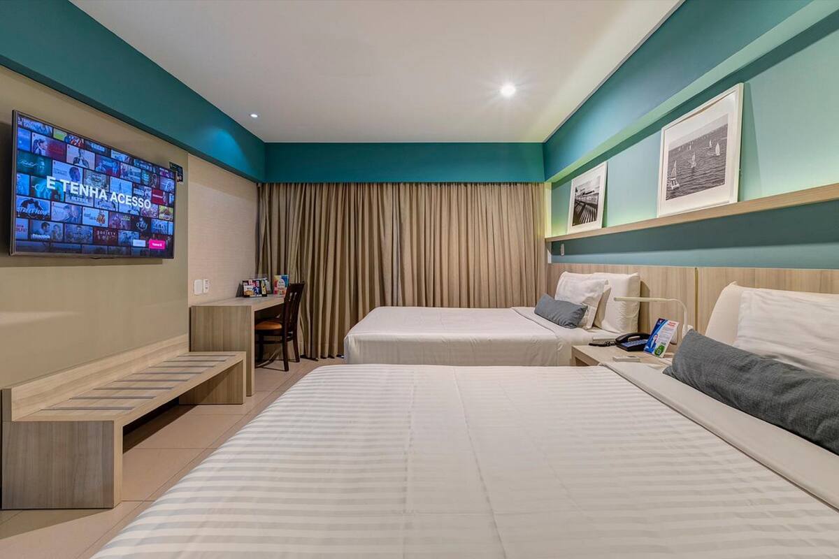 Ein Hotelzimmer mit zwei Betten, einem Schreibtisch, einem Fernseher und einem großen Fenster mit Vorhängen.