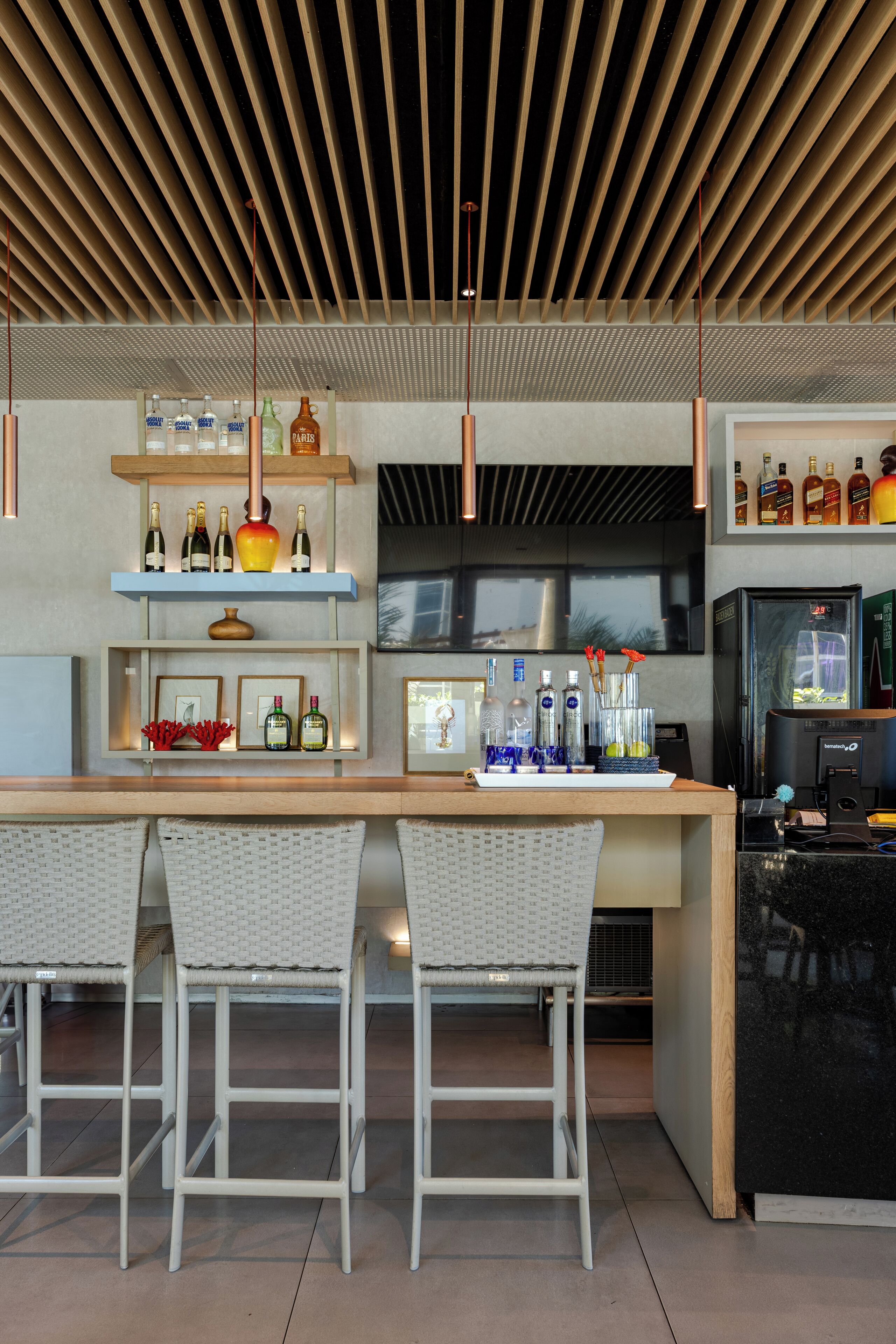 A modern bar area with a wooden ceiling, shelves with bottles, a refrigerator, and two white wicker chairs.