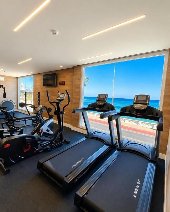 Une salle de sport équipée de tapis de course, de vélos d’appartement et d’appareils elliptiques, avec de grandes fenêtres offrant une vue sur l’océan.