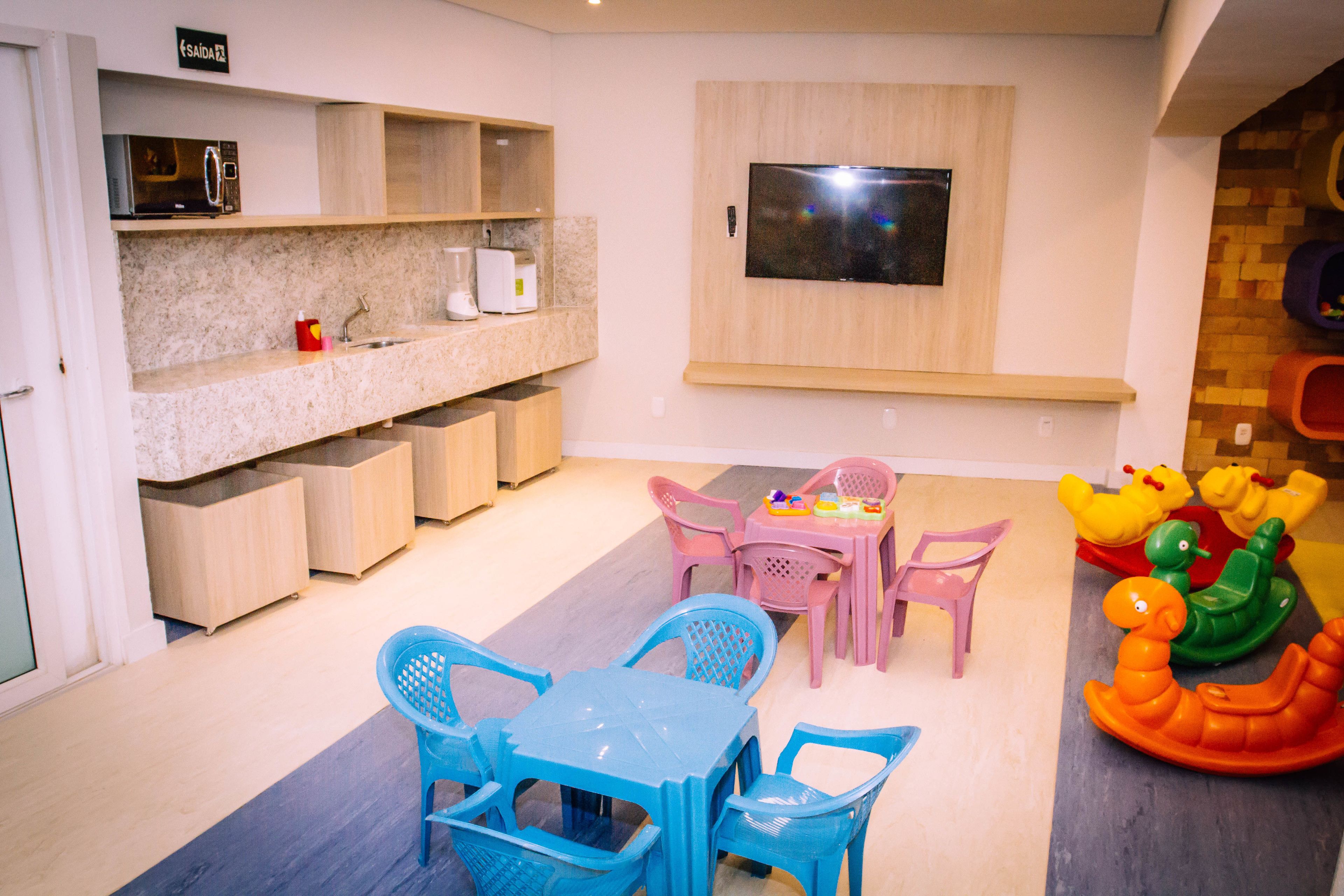 Een kinderspeelruimte met een tafel en stoelen, een aan de muur bevestigde televisie, en een speelmat met speelgoed.