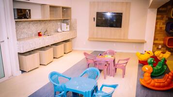 Ein Spielbereich für Kinder mit Tisch und Stühlen, einem an der Wand befestigten Fernseher und einer Spieledecke mit Spielzeug.