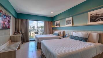 A hotel room with two beds, a small desk, a balcony with curtains, and framed pictures on the wall.