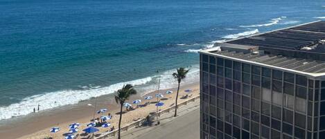 Vista costera con playa, sombrillas y un edificio de fachada de vidrio.