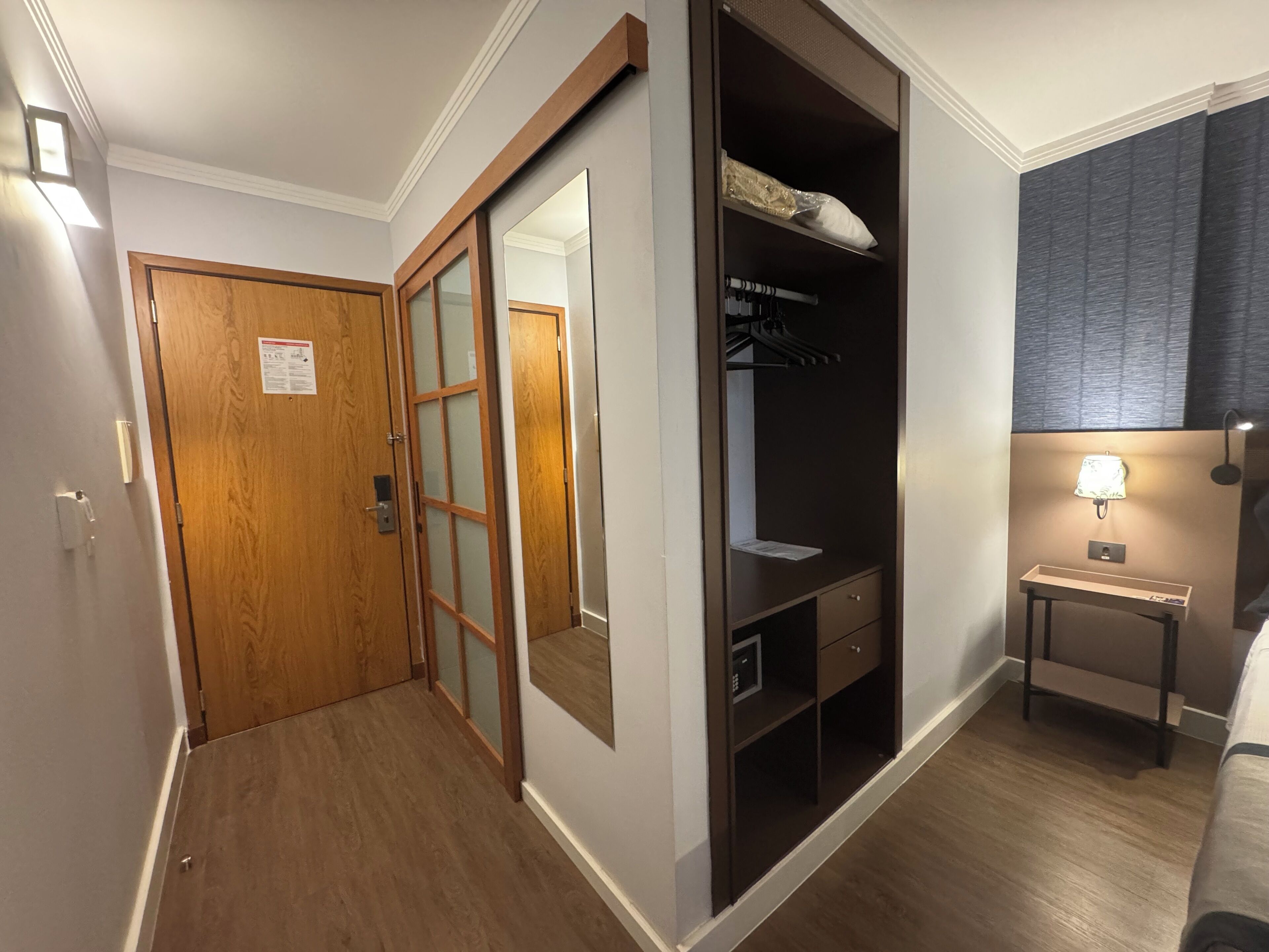 Quarto de hotel com porta de madeira, guarda-roupa com espelho, mesa de cabeceira com abajur e uma cama.