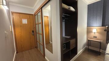 Quarto de hotel com porta de madeira, guarda-roupa com espelho, mesa de cabeceira com abajur e uma cama.