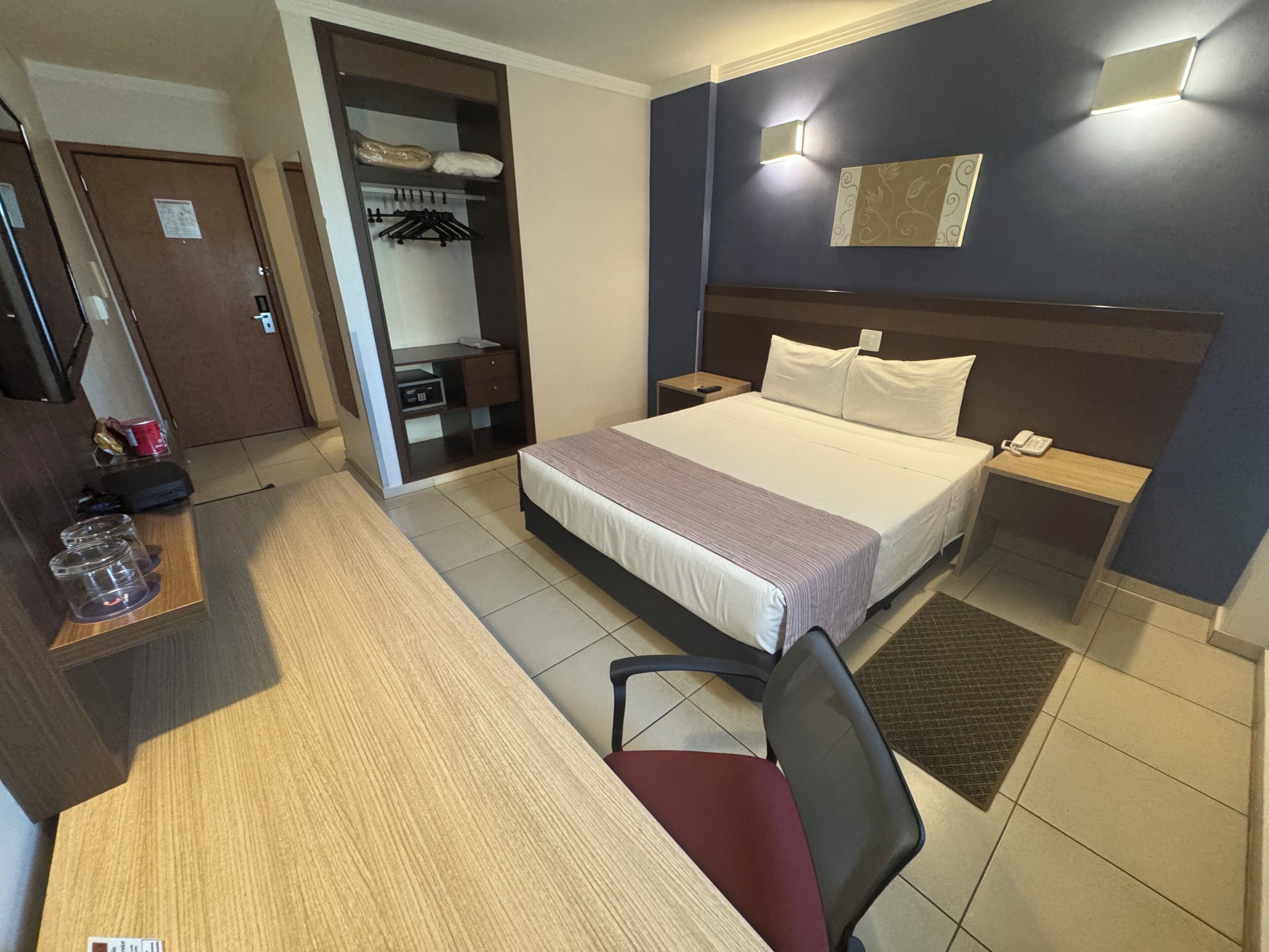 Quarto de hotel com cama, escrivaninha, cadeira, guarda-roupa e mesa de cabeceira com telefone.