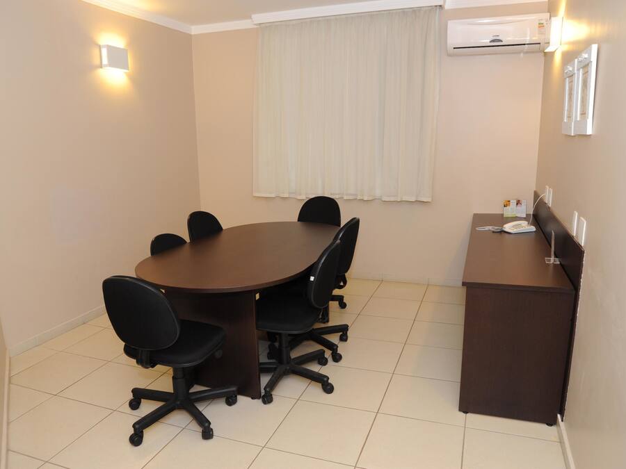 A conference room with a round table, six black office chairs, a brown desk, and a white air conditioning unit on the wall.
