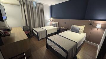 Una habitación de hotel con dos camas individuales, un escritorio con silla, televisor y ventana con cortinas.