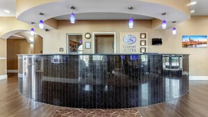 Una zona de recepción con un mostrador de cristal curvo, un cartel de 'Comfort Suites', un televisor montado y una pintura de un paisaje urbano en la pared.