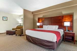Habitación de hotel con una cama grande, cabecera de madera, dos lámparas de noche, un sillón marrón, una mesita y suelo alfombrado.