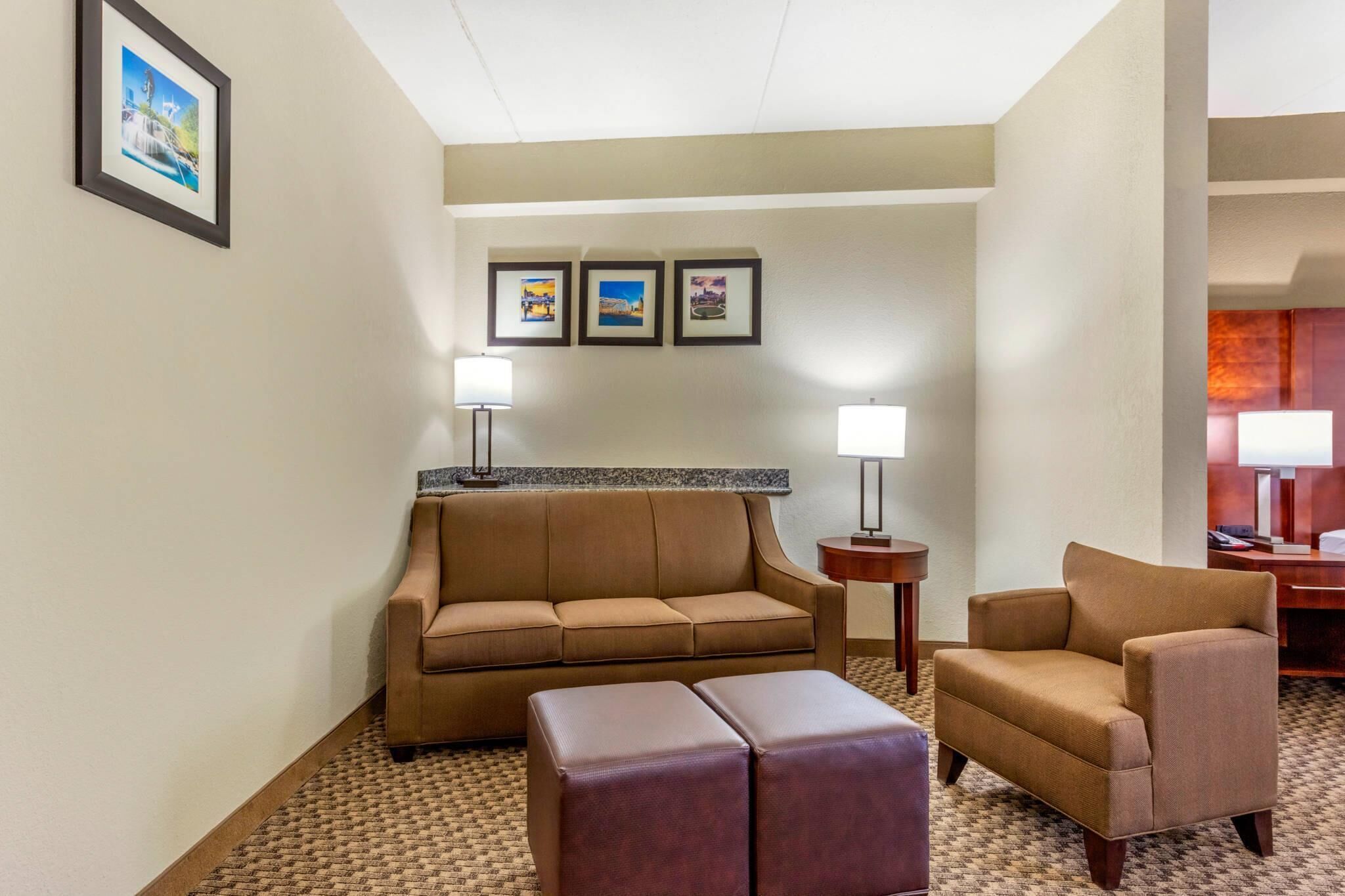 Quarto de hotel com um sofá castanho, uma poltrona a combinar, um pequeno banquinho e piso coberto por carpete. Há três quadros nas paredes e duas luminárias em mesas de cabeceira.