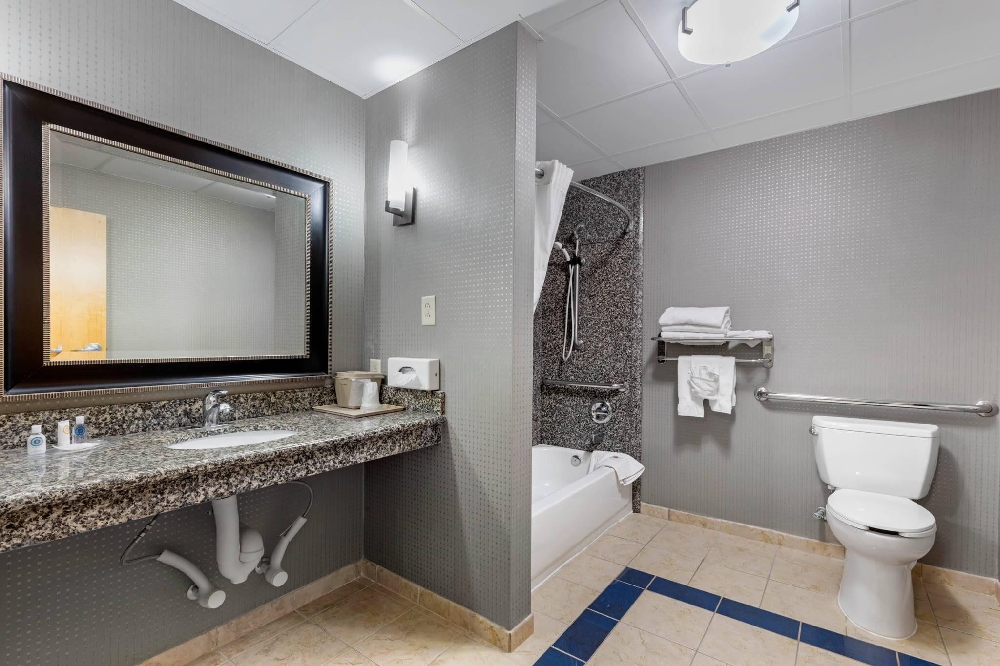 Un baño moderno con lavamanos de granito, un espejo grande, ducha con puerta de vidrio, inodoro blanco y barra de apoyo.