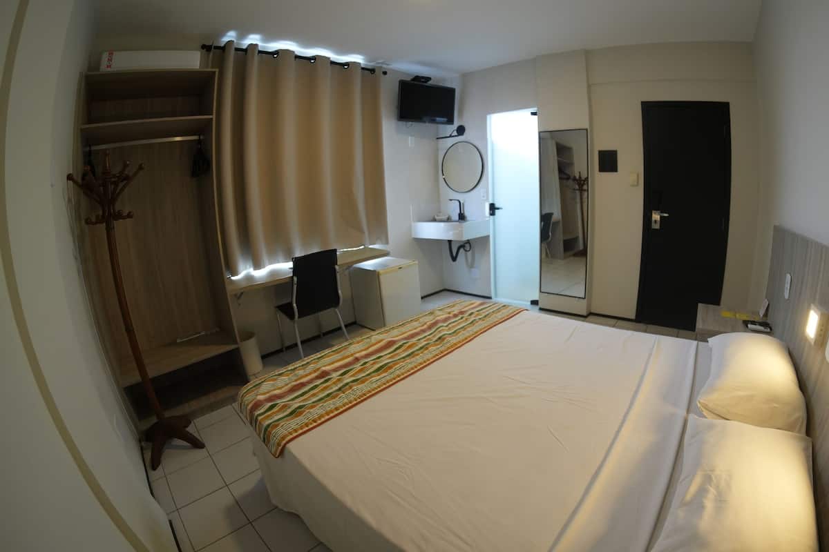 Una habitación de hotel con cama, escritorio, silla, armario, espejo, televisor y baño.