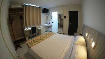 Quarto de hotel com cama, escrivaninha, cadeira, armário, espelho, televisão e banheiro.